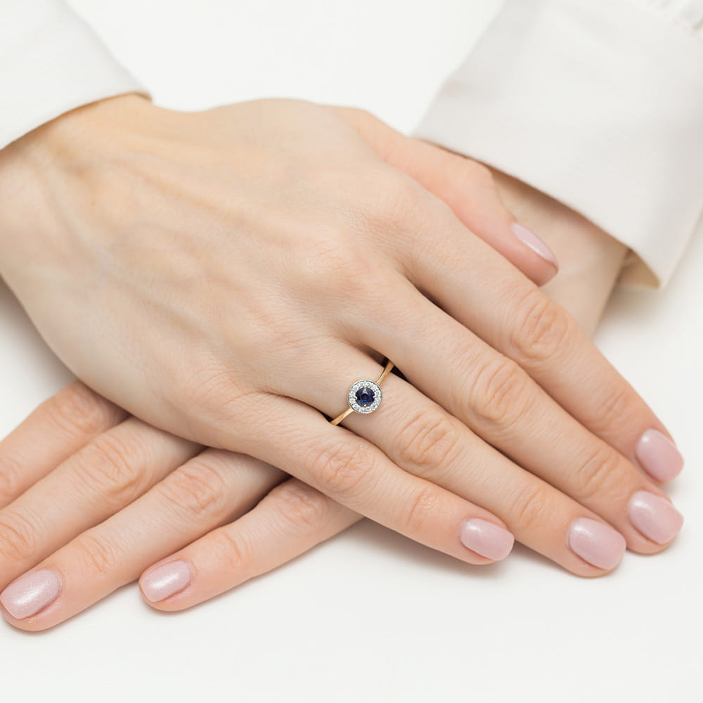 Die Hände einer Person zeigen einen eleganten Verlobungsring mit einem runden blauen Saphir und kleinen Diamanten auf einem schmalen goldenen Band.