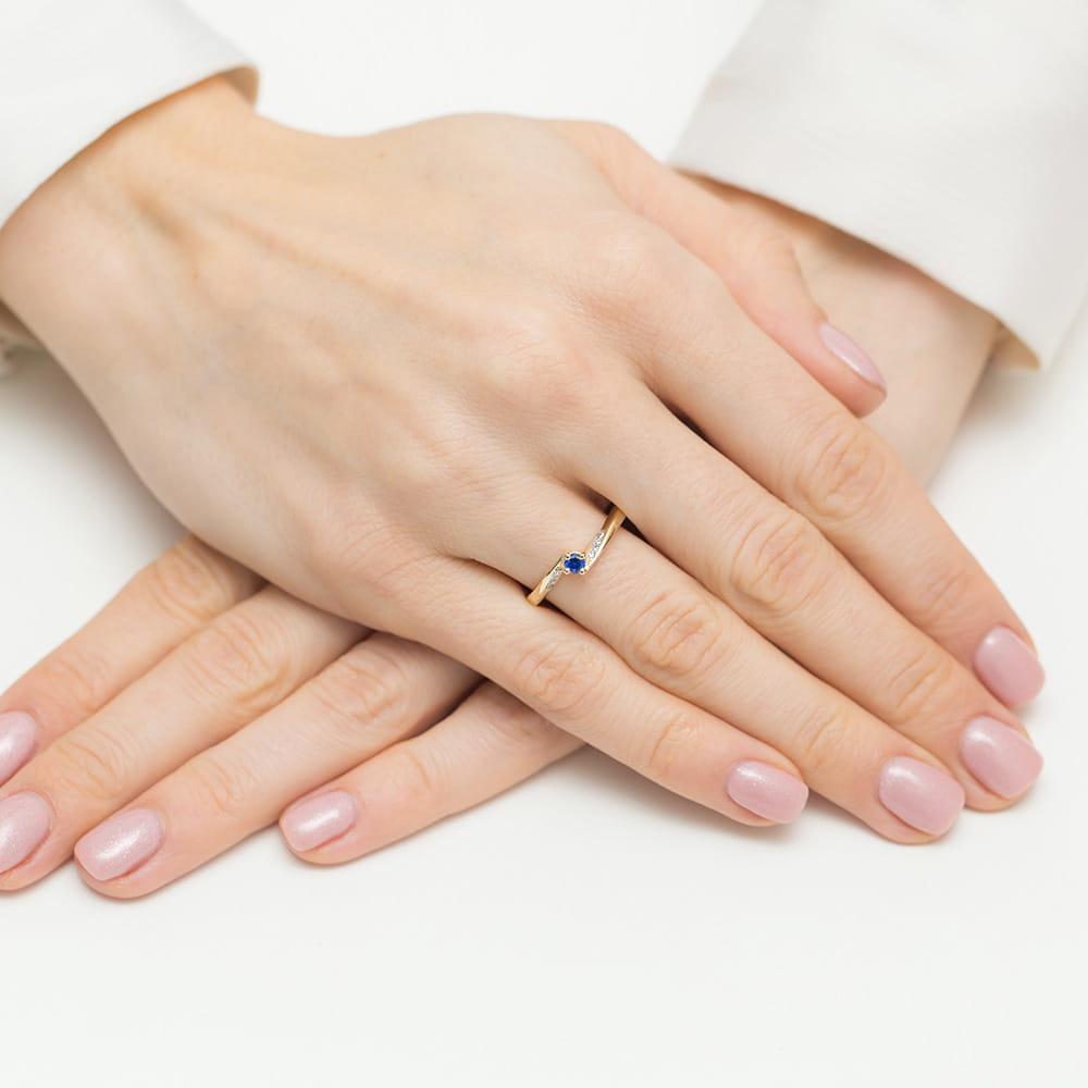 Hand mit einem eleganten Verlobungsring aus Gelbgold mit einem blauen Saphir und acht Diamanten. Der Ring sitzt perfekt am Finger und wirkt stilvoll und zeitlos.