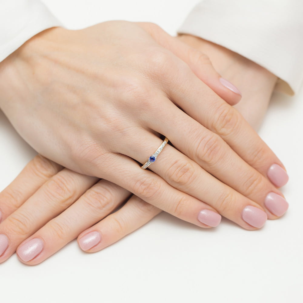 Ein eleganter Verlobungsring aus 585er Gold mit einem runden blauen Saphir als Hauptstein, flankiert von kleineren funkelnden Diamanten. Der Ring zeigt eine feine Handwerkskunst und eine klassische Designästhetik.