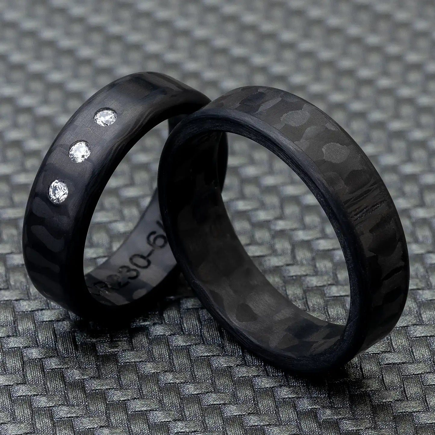 Zwei schwarze Carbon Trauringe auf einem strukturierten Hintergrund, einer mit drei Diamanten.