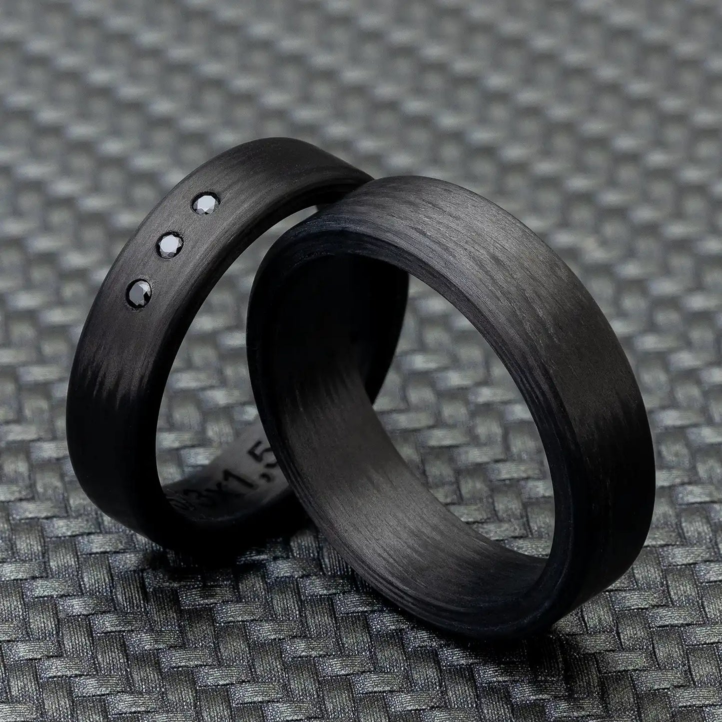 Zwei schwarze Carbon Eheringe auf einem grauen, strukturierten Hintergrund, einer verziert mit drei schwarzen Zirkonia.