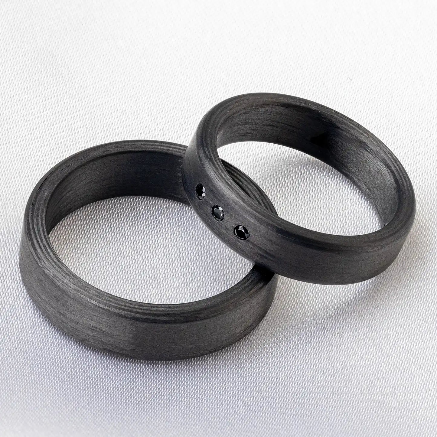 Zwei Carbon Trauringe auf einem hellen Stoffhintergrund, einer verziert mit drei schwarzen Zirkonia.