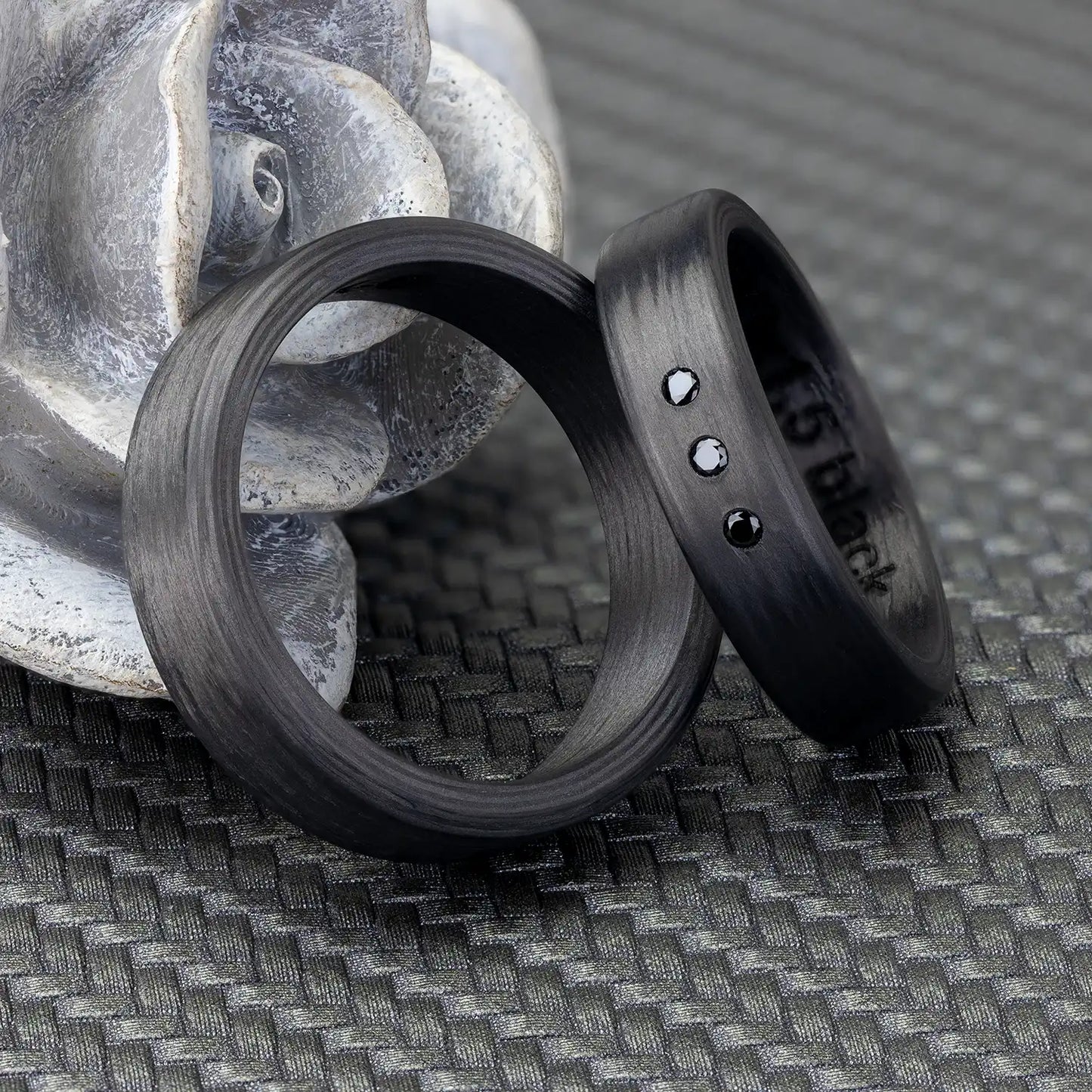 Zwei Carbon Eheringe vor einer grauen Rosenfigur, einer verziert mit drei schwarzen Zirkonia.