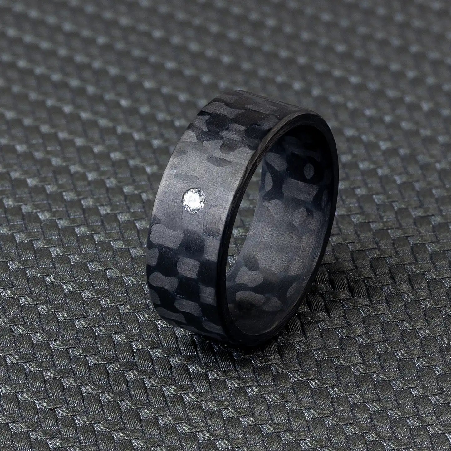 Schwarzer Karbonfaser-Ring mit Diamant oder Zirkonia auf strukturiertem Hintergrund.