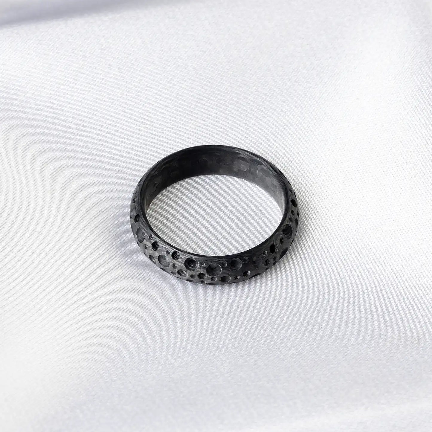 Ein schwarzer Carbonfaser-Ring mit mondkraterähnlicher Textur, auf einer weißen Oberfläche liegend.