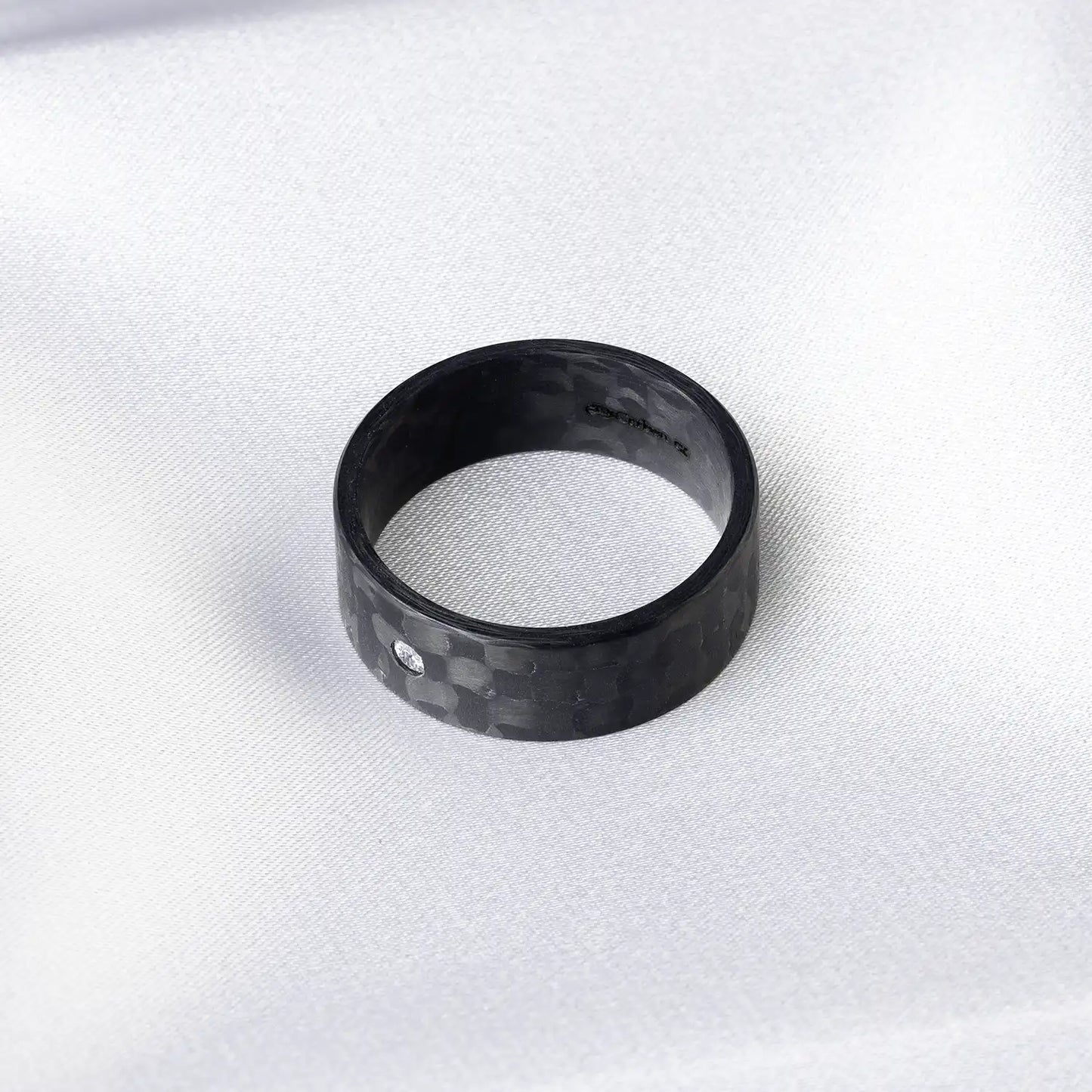 Schwarzer Karbonfaser-Ring mit Diamant oder Zirkonia auf weißem Stoffhintergrund.