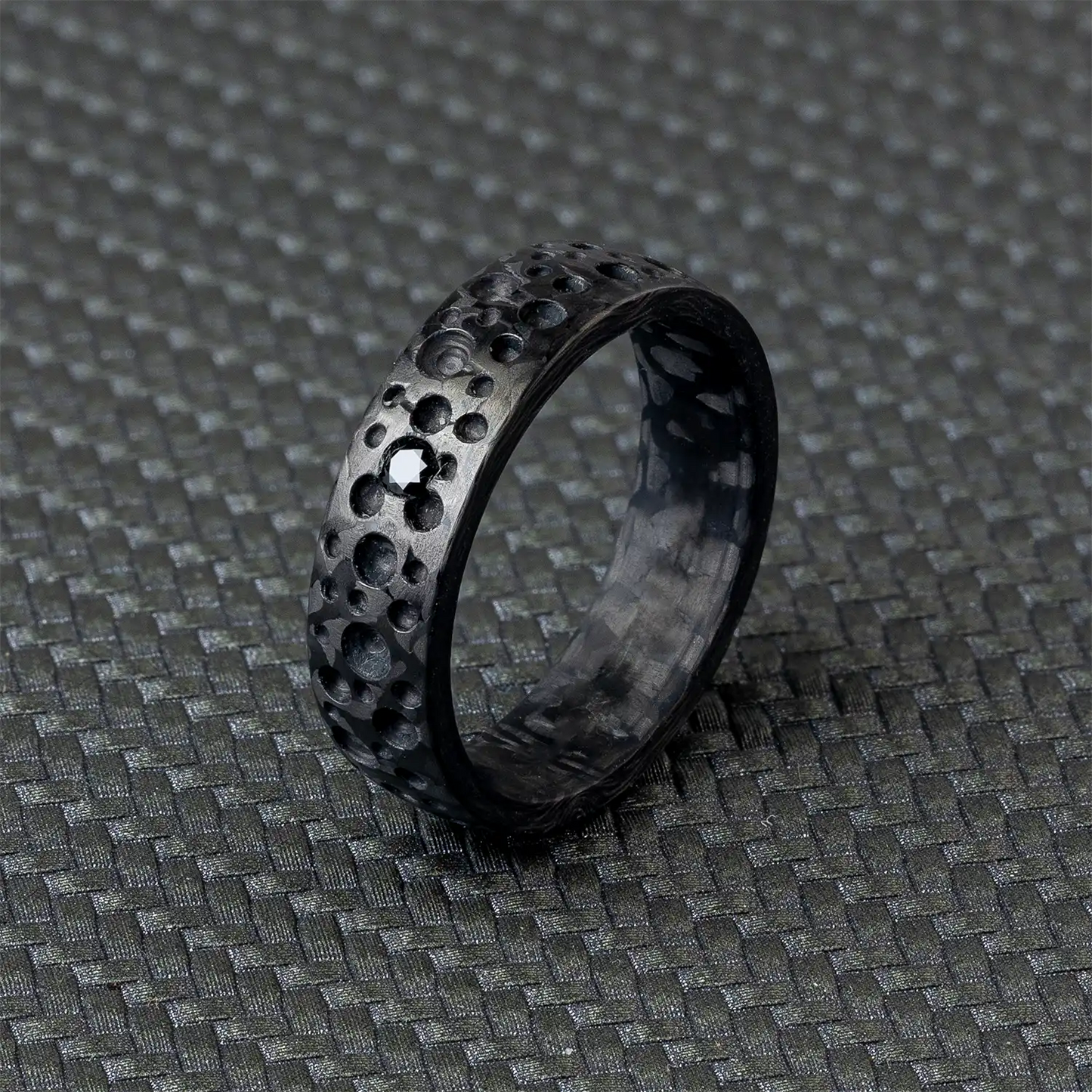 Schwarzkarbon-Ring mit texturierter Oberfläche, die an Mondkrater erinnert, aus Froschperspektive auf schwarzem Hintergrund.