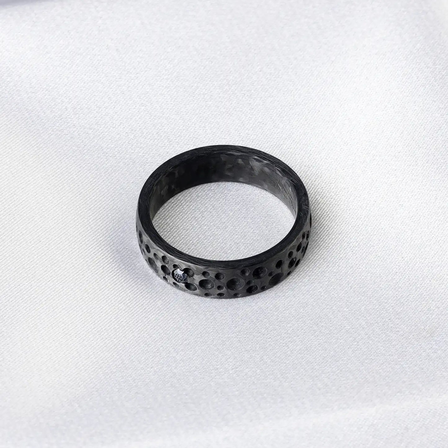 Schwarzkarbon-Ring mit Mondkratertextur und einem schwarzen Diamanten oder Zirkonia auf weißem Stoffhintergrund.