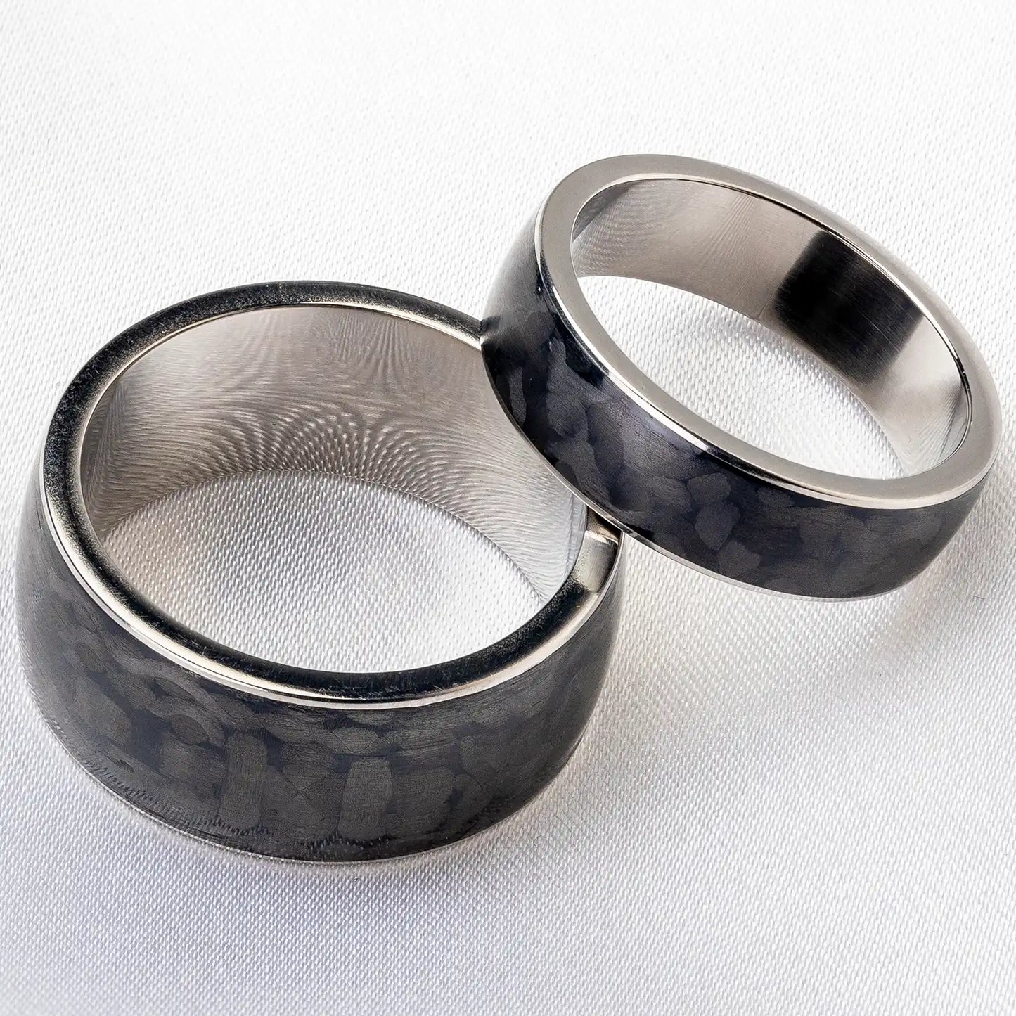 Schwarze Eheringe aus Titan und Karbonfaser auf einem weißen Stoffhintergrund, ein breiter Ring für Männer und ein schmaler Ring für Frauen.