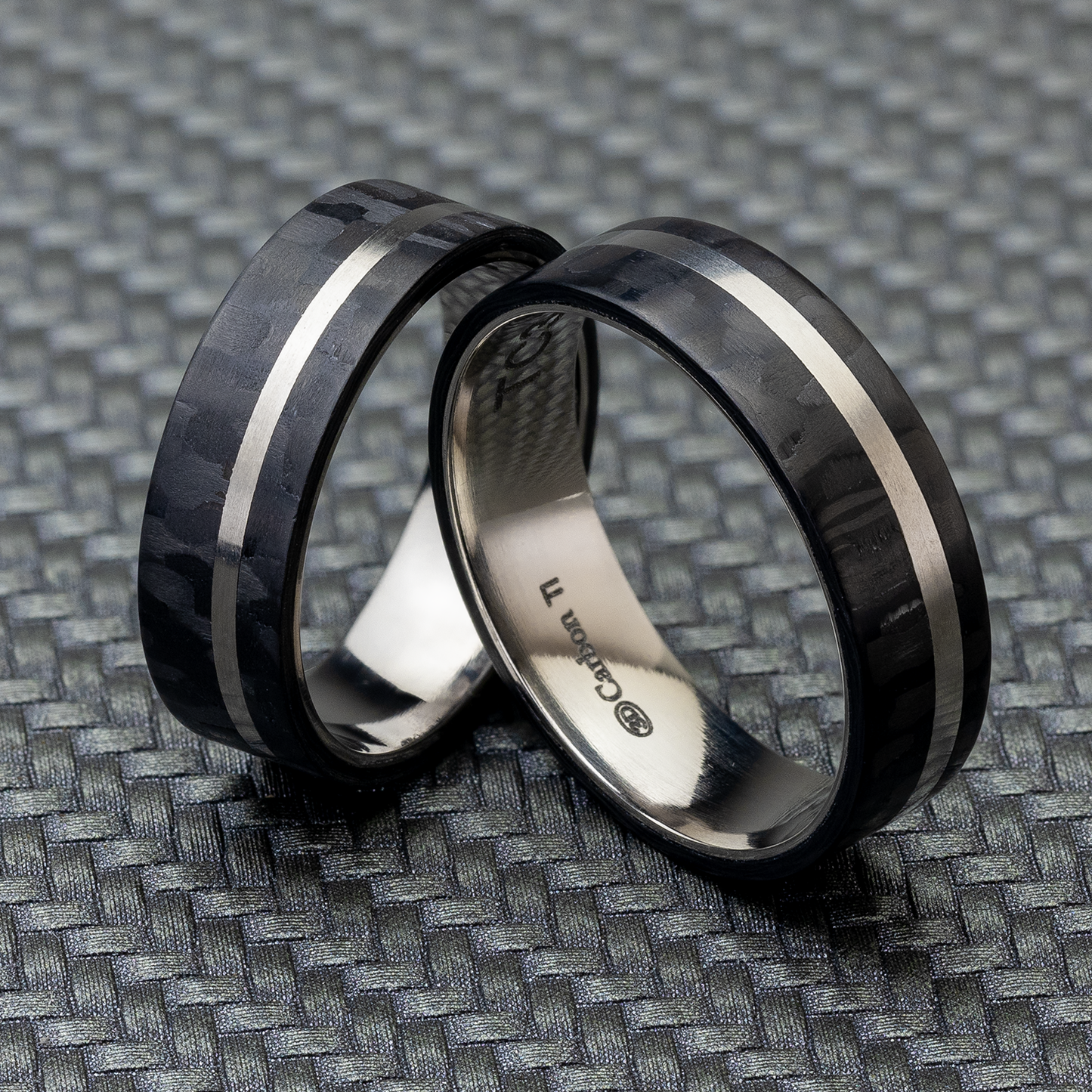 Zwei schwarze Carbon Partnerringe mit Titan und einer Breite von 6 mm, stehend auf einer gemusterten Oberfläche.