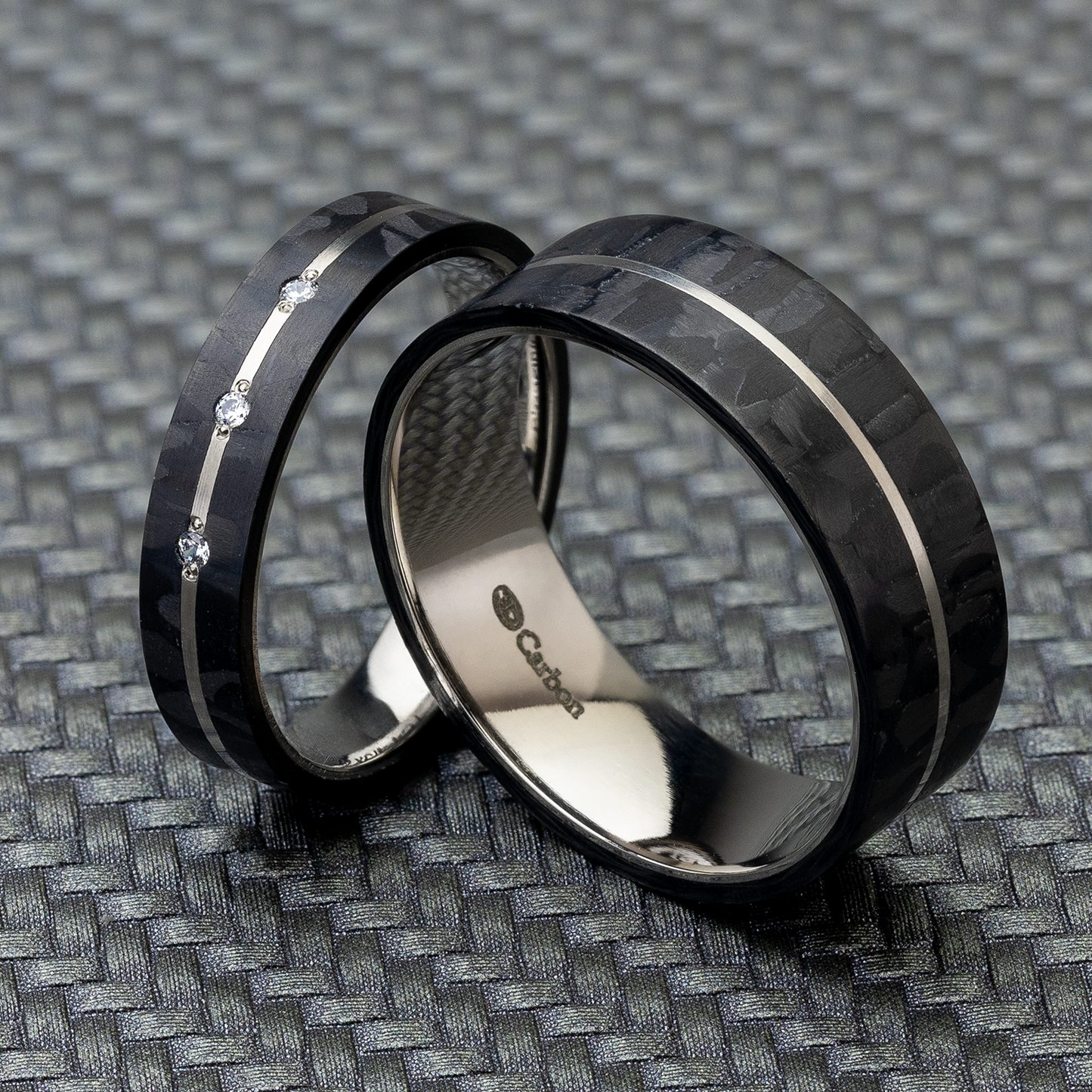 Schwarze Partnerringe aus Carbon und Titan auf einem gemusterten Hintergrund. Der Damenring hat 3 Diamanten, der Herrenring ist schlicht.