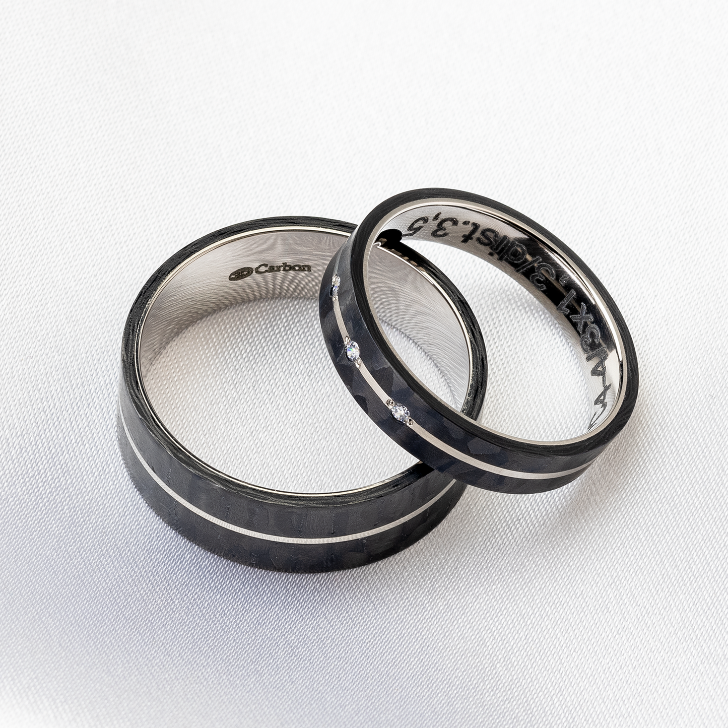 Schwarze Carbon-Trauringe auf weißem Stoff. Der Damenring mit 3 Diamanten und der Herrenring nebeneinander liegend.
