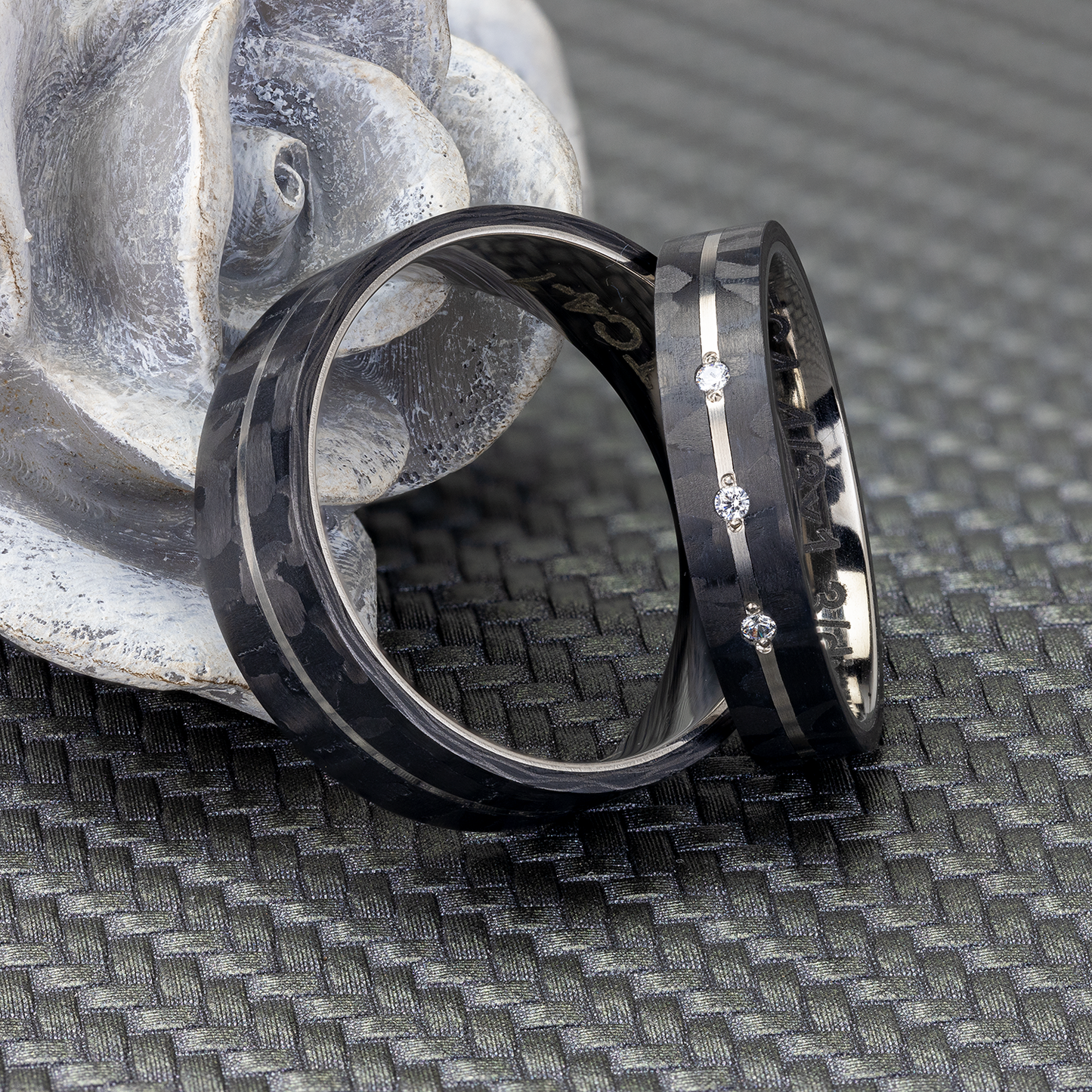 Zwei Carbon Eheringe aus Carbon und Titan, einer mit drei Zirkonias, auf einer grauen Rose liegend.