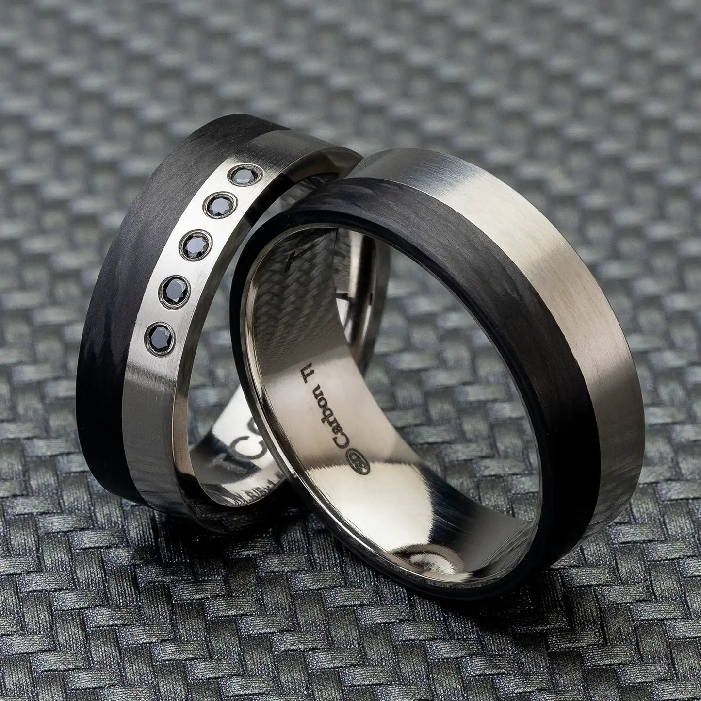 Zwei Carbon Partnerringe und Titan, einer mit fünf schwarzen Diamanten, auf einer strukturierten Oberfläche.