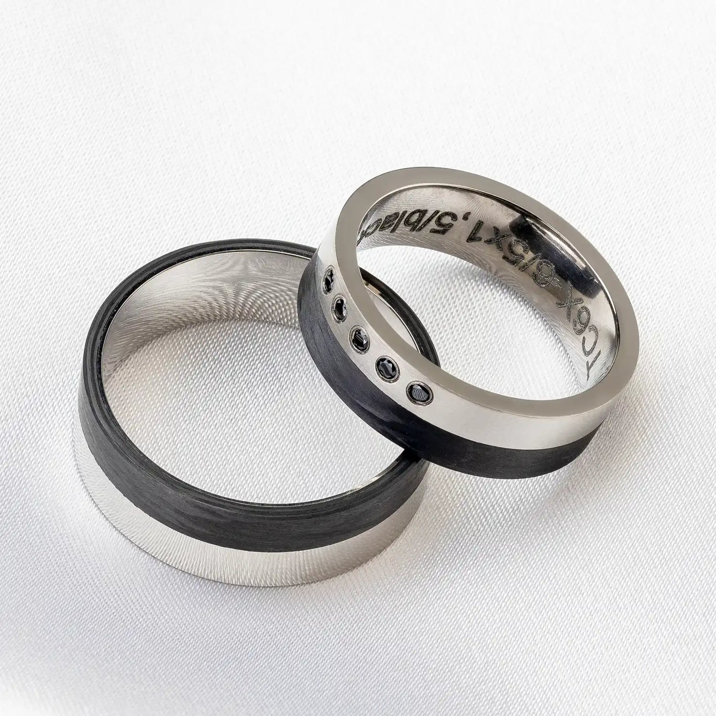 Zwei Carbon Eheringe, einer davon mit fünf schwarzen Zirkonia, auf einem weißen Hintergrund.
