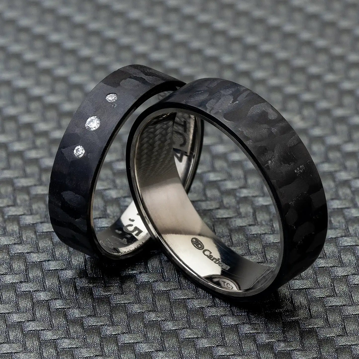 Zwei schwarze Carbon und Titan Ringe, nebeneinander auf einer strukturierten Oberfläche platziert. Der Damenring ist mit drei Zirkonia-Steinen besetzt.