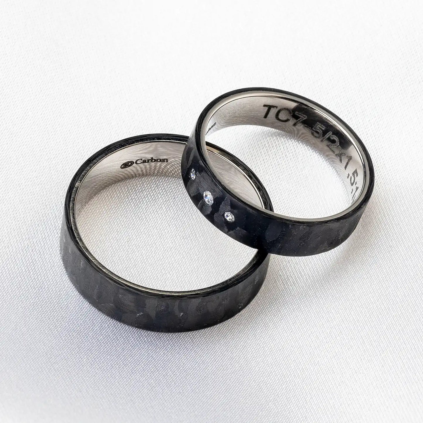 Zwei schwarze Carbon und Titan Trauringe auf einer weißen Oberfläche, der Damenring hat drei kleine Diamanten und der Herrenring ist schlicht.