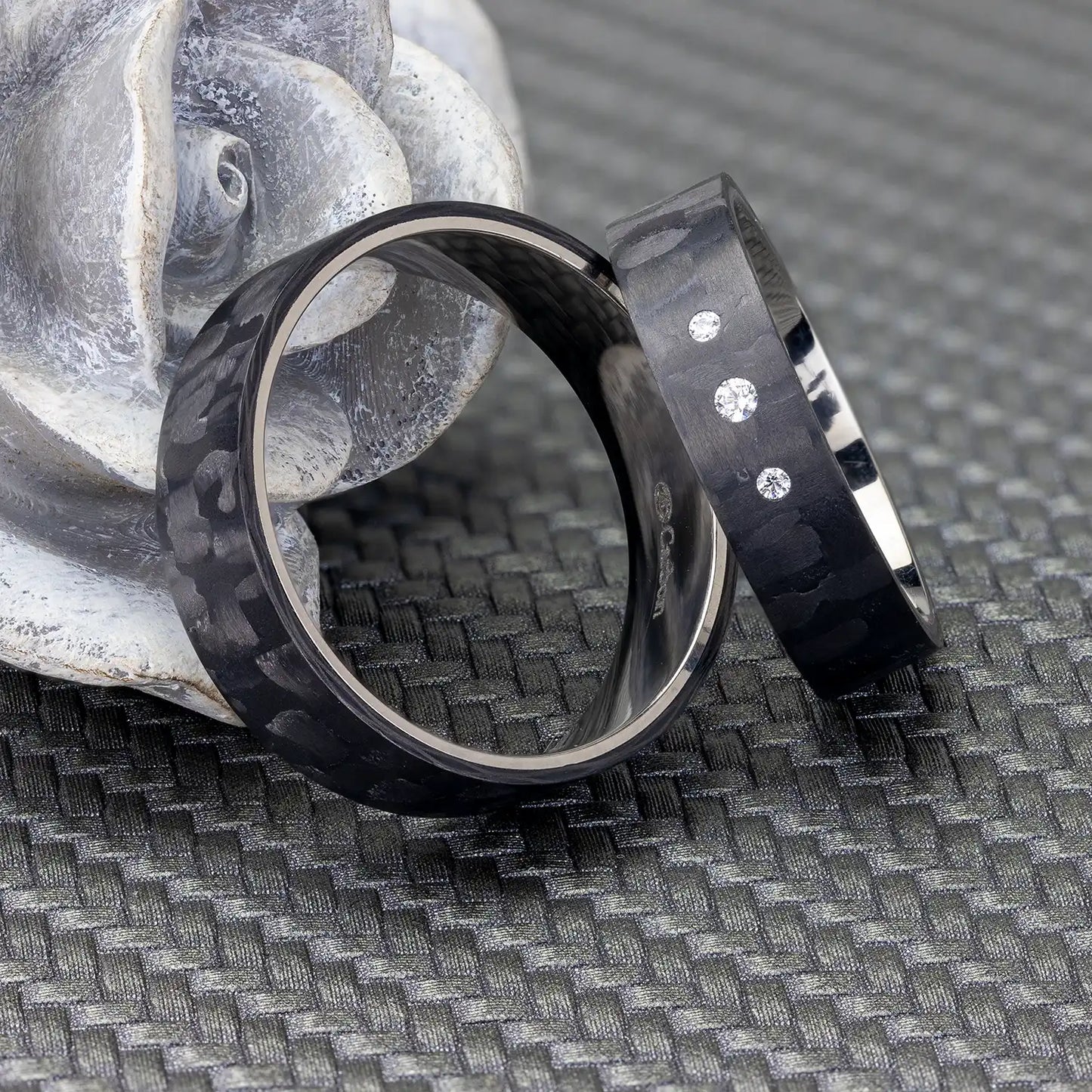 Zwei schwarze Carbon und Titan Partnerringe, elegant platziert vor einer grauen, rosafarbenen Dekoration. Der Damenring ist mit drei Zirkonia-Steinen verziert.
