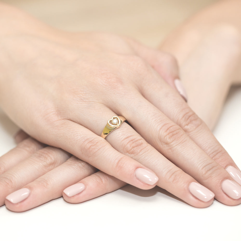 Goldener Verlobungsring mit einem herzförmigen Diamanten, getragen an der Hand einer Person. Die Fingernägel sind gepflegt und hellrosa lackiert.