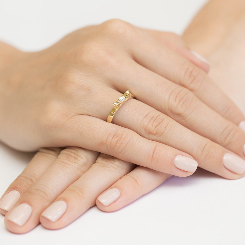 Eine Nahaufnahme von zwei Händen, die eine zarte Goldring mit einer Schleife und einem kleinen Diamanten tragen. Der Ring ist elegant und perfekt für eine Verlobung.