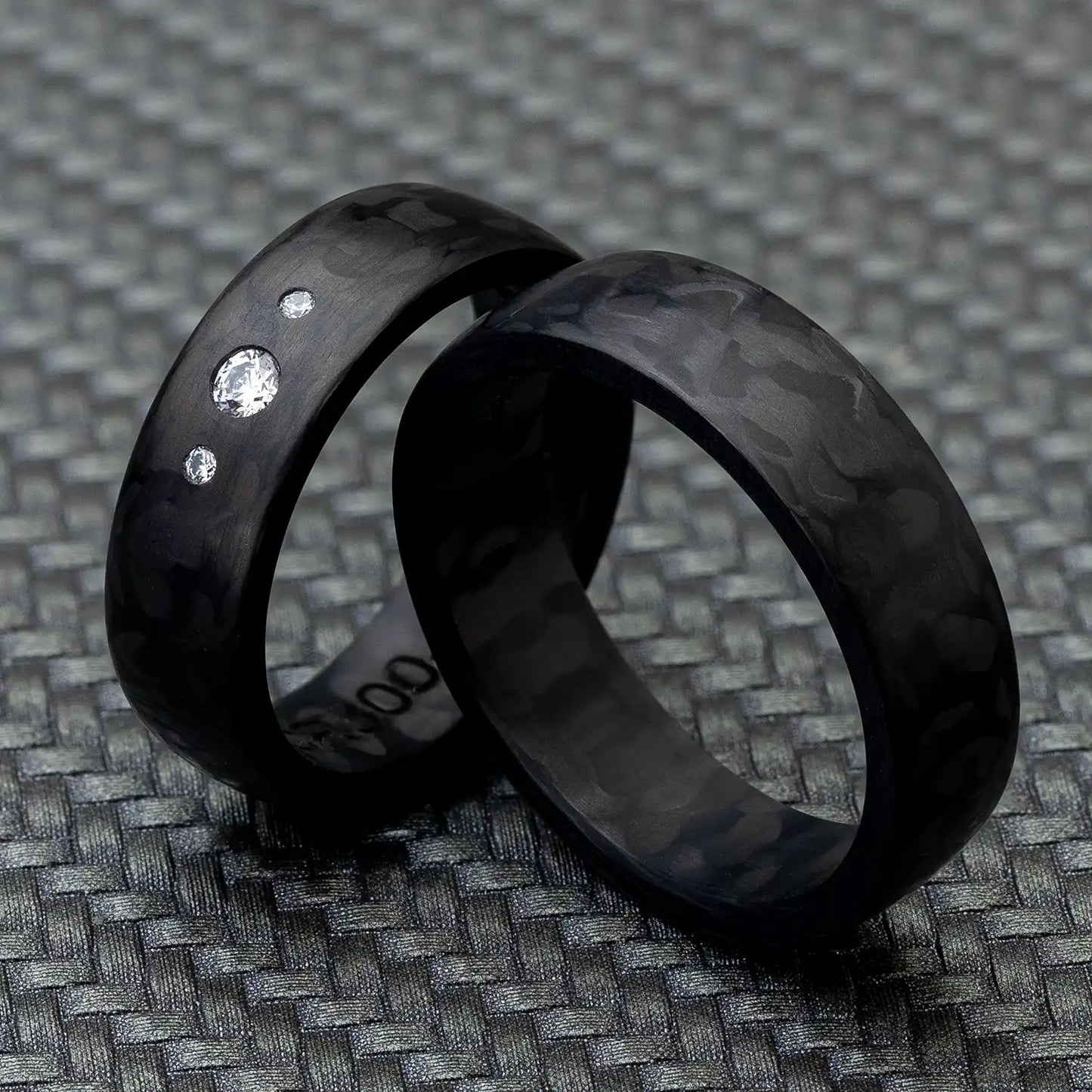 Schwarze Carbon Eheringe, einer mit 3 Diamanten, auf einem strukturierten, dunklen Hintergrund.