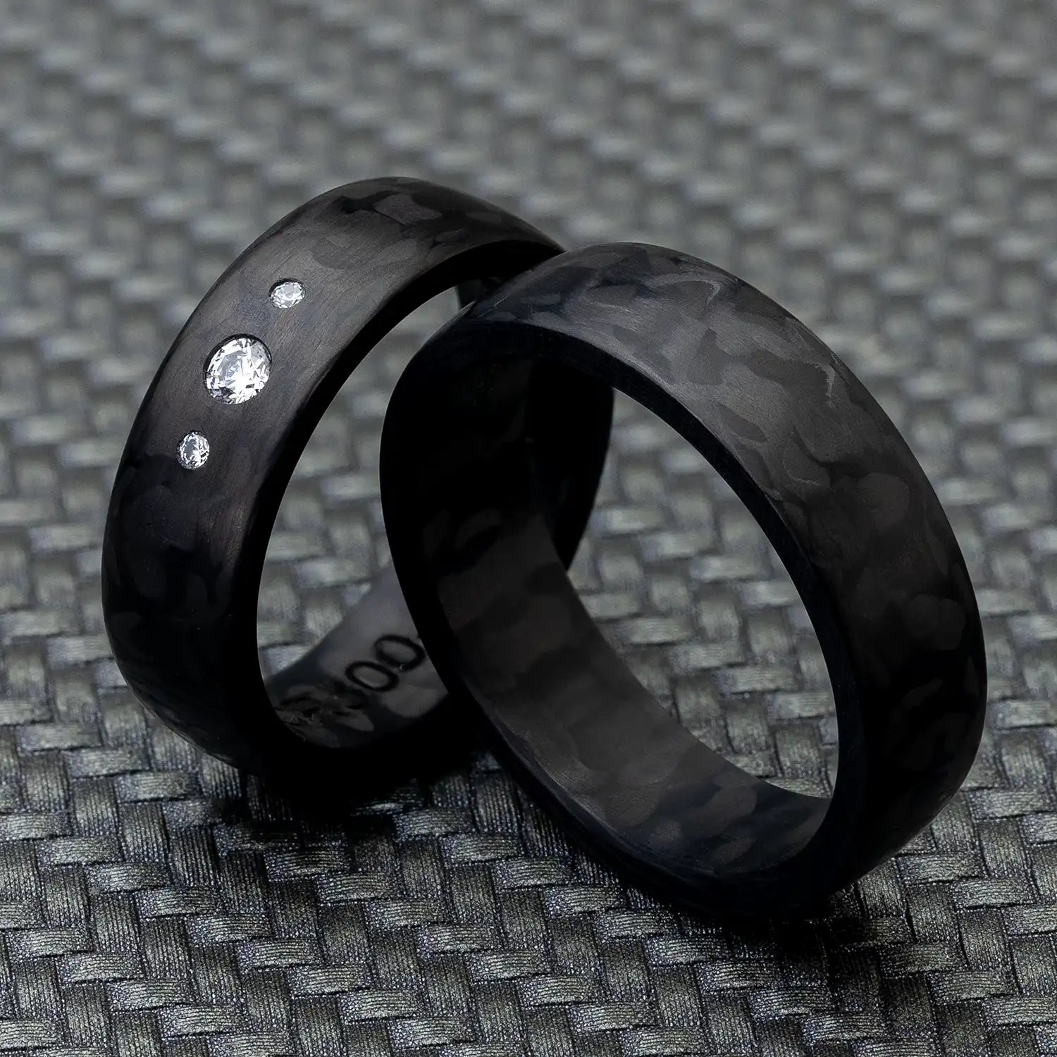 Schwarze Carbon Eheringe, einer mit 3 Diamanten, auf einem strukturierten, dunklen Hintergrund.