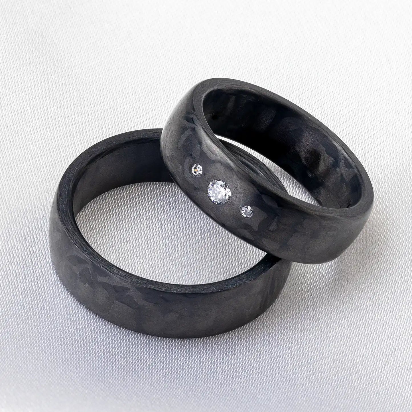 Zwei schwarze Carbon Eheringe, einer davon mit 3 Diamanten, auf einem hellen Stoffhintergrund.