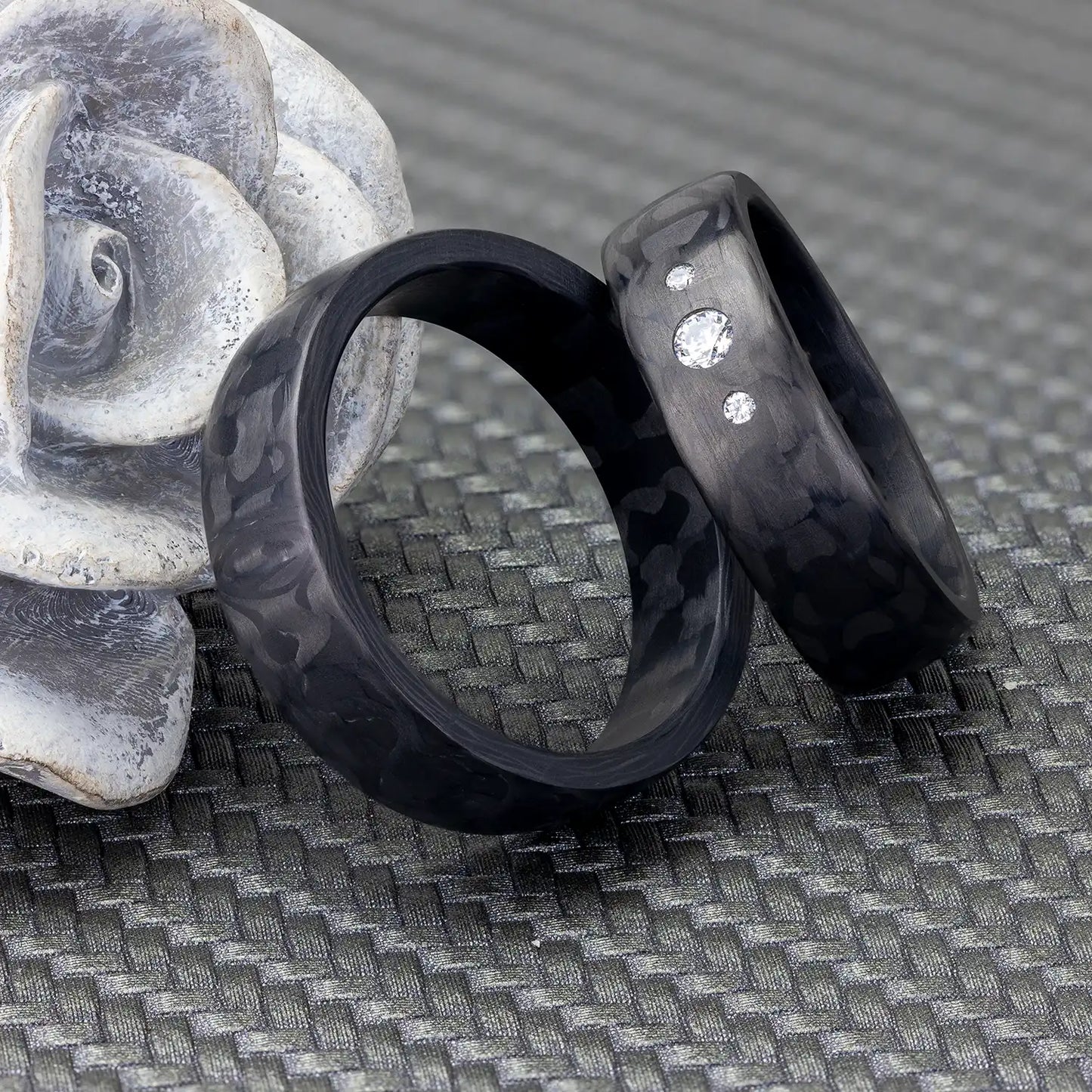 Schwarze Carbon Eheringe mit 3 Diamanten neben einer weißen Blumenfigur auf einem gewebten Hintergrund.