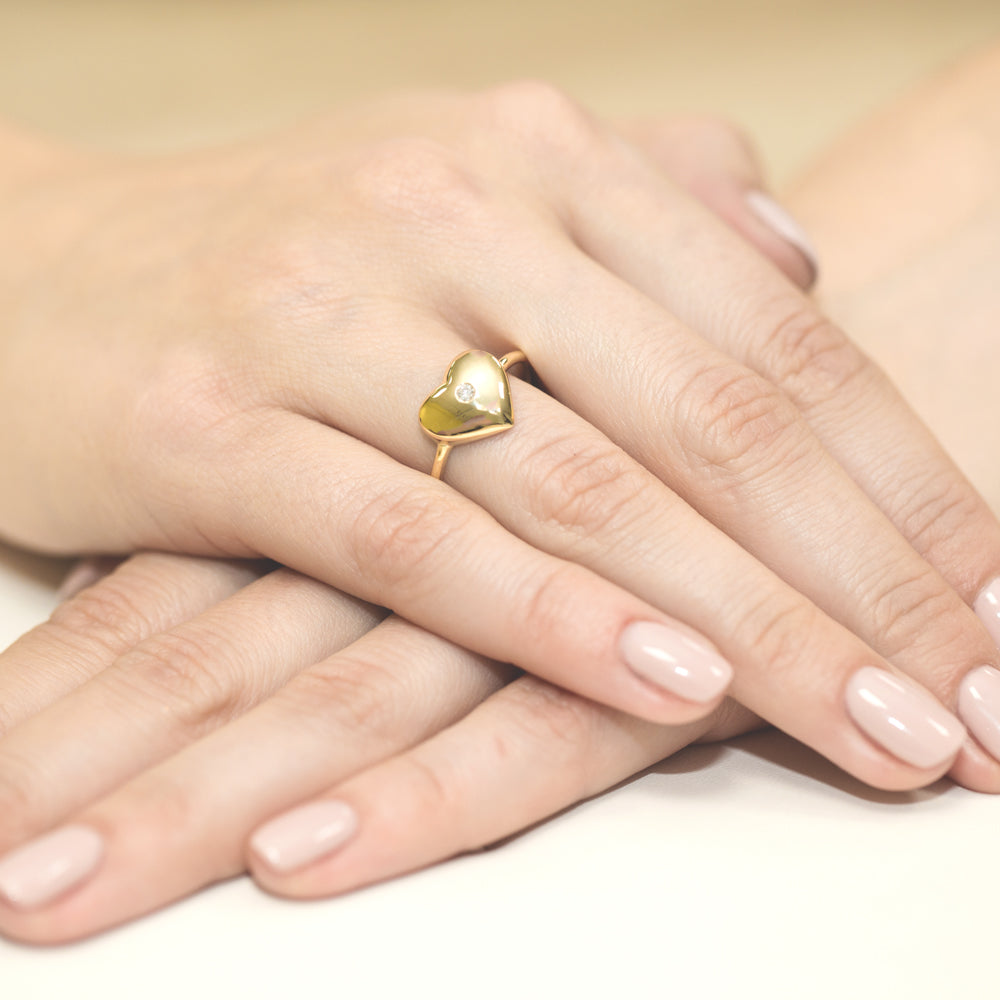 Ein Goldring mit einem herzförmigen Design und einem kleinen, zentralen Diamanten. Der Ring ist an einer Hand getragen und zeigt elegante, gepflegte Fingernägel.