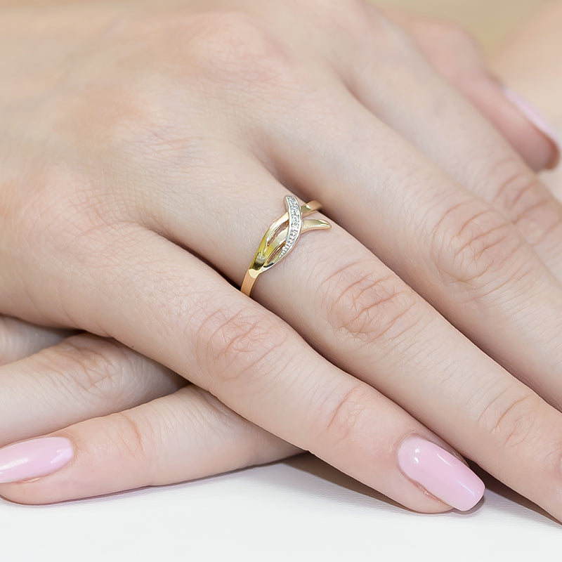 Ein eleganter Verlobungsring aus Gelbgold 585 mit einem funkelnden Diamanten von 0,02ct auf einer Hand. Der Ring hat ein modernes, geschwungenes Design und die Nägel sind rosa lackiert.