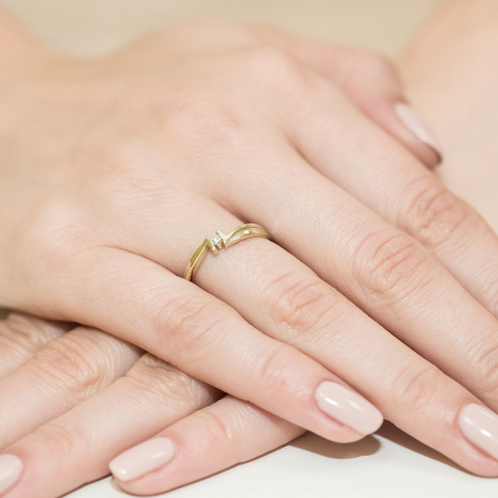 Dieser Verlobungsring aus Gelbgold hat eine geschwungene Form und ist mit einem kleinen, funkelnden Diamanten besetzt. Der Ring ist auf einer Hand getragen, was seine Eleganz und zarte Verarbeitung betont.